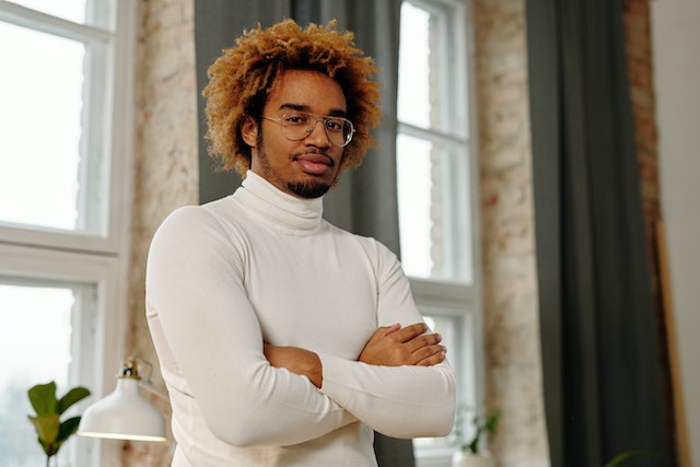 Man met donkere huidskleur, witte turtle neck, pilot glasses en een geblondeerd afrokapsel

